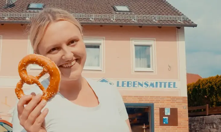 Christina Schönberger lächelt und hält eine Breze in der Hand. Sie steht vor einem Haus mit der Aufschrift „Lebensmittel“.